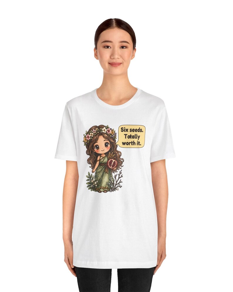 May include: A white t-shirt featuring a graphic of a cartoon character with long brown hair, a flower crown, and a green dress, holding a pomegranate. A yellow speech bubble next to the character contains the text: Six seeds. Totally worth it.