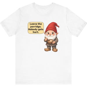 May include: A white t-shirt featuring a graphic of a gnome wearing a red hat and brown clothing, holding a wooden bowl and spoon. A speech bubble next to the gnome contains the text: Leave the porridge. Nobody gets hurt.