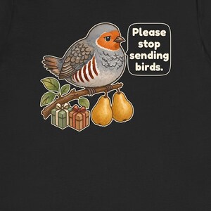 K&ouml;nnte beinhalten: Ein schwarzes T-Shirt mit einer Grafik eines grauen und orangefarbenen Vogels, der auf einem Ast mit zwei gelben Birnen und zwei kleinen Geschenkkartons sitzt. Eine Sprechblase neben dem Vogel enth&auml;lt den wei&szlig;en Text: Please stop sending birds.