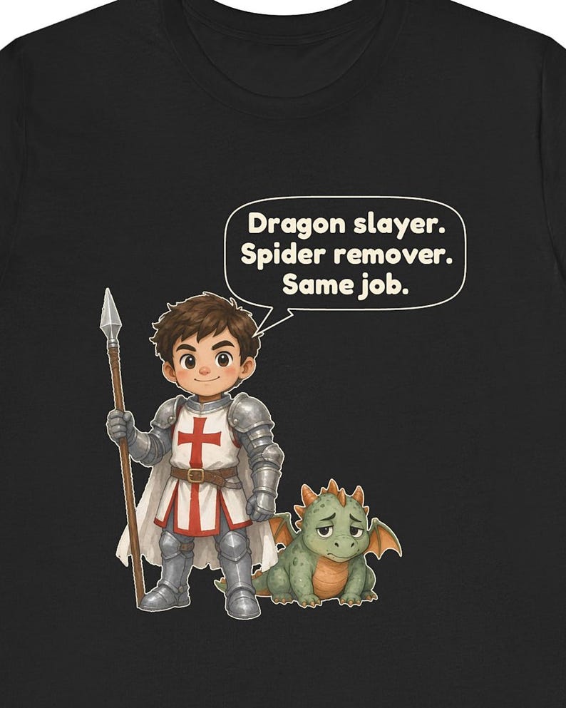 K&ouml;nnte beinhalten: Ein schwarzes T-Shirt mit einem Cartoon-Ritter in silberner R&uuml;stung, der eine wei&szlig;e Tunika mit rotem Kreuz tr&auml;gt und einen Speer h&auml;lt. Neben dem Ritter ist ein kleiner gr&uuml;ner Drache. Eine Sprechblase enth&auml;lt den Text: Dragon slayer. Spider remover. Same job.