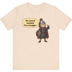 May include: A light beige t-shirt featuring a cartoon illustration of a warrior in chainmail and a helmet with a small animal on top. The warrior is flexing one arm and holding a round shield. A speech bubble contains the text: No sword needed. I'm a hugger.