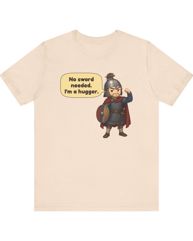May include: A light beige t-shirt featuring a cartoon illustration of a warrior in chainmail and a helmet with a small animal on top. The warrior is flexing one arm and holding a round shield. A speech bubble contains the text: No sword needed. I'm a hugger.