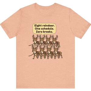 K&ouml;nnte beinhalten: Ein pfirsichfarbenes Kurzarm-T-Shirt mit einer Grafik von acht braunen Rentieren in zwei Reihen. &Uuml;ber den Rentieren befindet sich eine gelbe Sprechblase mit dem schwarzen Text: Eight reindeer. One schedule. Zero breaks.
