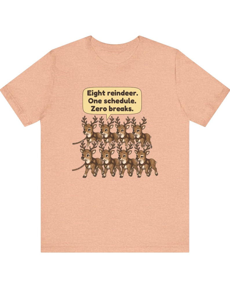 K&ouml;nnte beinhalten: Ein pfirsichfarbenes Kurzarm-T-Shirt mit einer Grafik von acht braunen Rentieren in zwei Reihen. &Uuml;ber den Rentieren befindet sich eine gelbe Sprechblase mit dem schwarzen Text: Eight reindeer. One schedule. Zero breaks.