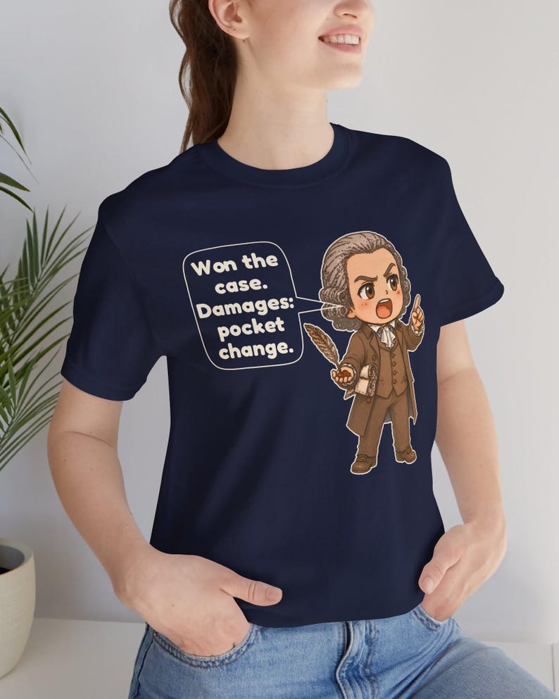 May include: A navy blue t-shirt featuring a cartoon illustration of a historical figure in a brown suit holding a quill. A speech bubble contains the text: Won the case. Damages: pocket change. The design is centered on the front of the shirt.