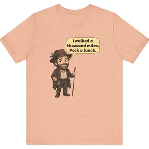 May include: A peach-colored short-sleeve t-shirt featuring a cartoon illustration of a bearded hiker wearing a brown hat with a pine sprig, a brown coat, and holding a wooden walking stick. A speech bubble contains the text: I walked a thousand miles. Pack a lunch.