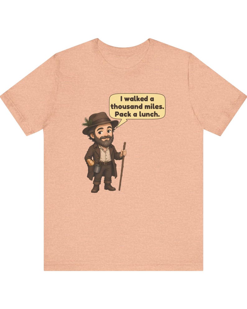 May include: A peach-colored short-sleeve t-shirt featuring a cartoon illustration of a bearded hiker wearing a brown hat with a pine sprig, a brown coat, and holding a wooden walking stick. A speech bubble contains the text: I walked a thousand miles. Pack a lunch.