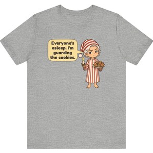 Op de afbeelding: Een grijs gem&ecirc;leerd t-shirt met een cartoonillustratie van een persoon in een rood-wit gestreepte pyjama en een bijpassende slaapmuts. De persoon houdt een brandende kaars en een bord met koekjes vast, met een tekstballon: Everyone's asleep. I'm guarding the cookies.