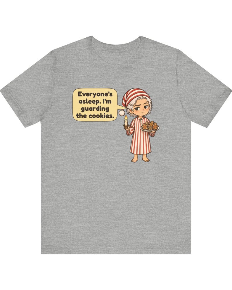 Op de afbeelding: Een grijs gem&ecirc;leerd t-shirt met een cartoonillustratie van een persoon in een rood-wit gestreepte pyjama en een bijpassende slaapmuts. De persoon houdt een brandende kaars en een bord met koekjes vast, met een tekstballon: Everyone's asleep. I'm guarding the cookies.