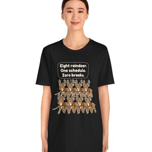 K&ouml;nnte beinhalten: Ein schwarzes Kurzarm-T-Shirt mit einer Grafik von acht braunen Rentieren, die in zwei Reihen angeordnet sind. &Uuml;ber den Rentieren befindet sich eine wei&szlig;e Sprechblase mit dem Text: Eight reindeer. One schedule. Zero breaks.