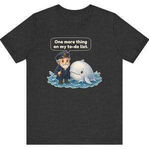 May include: A dark heather gray t-shirt featuring a graphic of a cartoon sea captain with a white beard and a white whale in blue water. A speech bubble above them contains the text, One more thing on my to-do list. The design is centered.