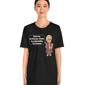Pu&ograve; includere: Una t-shirt nera con un'illustrazione a cartoni animati di un uomo anziano con gli occhiali, che indossa un cappotto viola e tiene in mano una penna e una pergamena. Un fumetto contiene il testo: Hebrew professor. Poet. Accidentally Christmas.