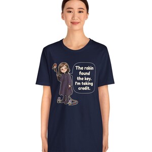 May include: A navy blue t-shirt featuring a graphic of a person in a dark coat holding a large key with a robin perched on it. A speech bubble contains the text: The robin found the key. I'm taking credit. The design includes small flowers.