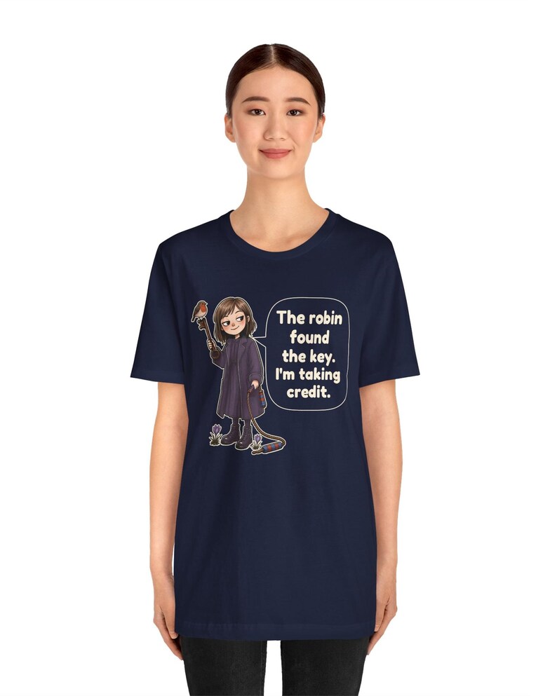 May include: A navy blue t-shirt featuring a graphic of a person in a dark coat holding a large key with a robin perched on it. A speech bubble contains the text: The robin found the key. I'm taking credit. The design includes small flowers.