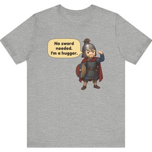 May include: A heather gray t-shirt featuring a cartoon illustration of a warrior in chainmail and a helmet with a small animal on top. The warrior holds a round shield and has a speech bubble that reads, No sword needed. I'm a hugger.