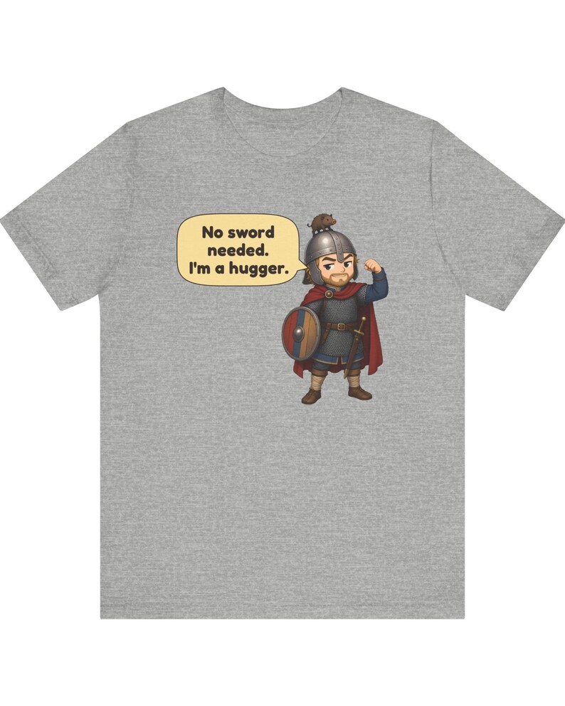 May include: A heather gray t-shirt featuring a cartoon illustration of a warrior in chainmail and a helmet with a small animal on top. The warrior holds a round shield and has a speech bubble that reads, No sword needed. I'm a hugger.