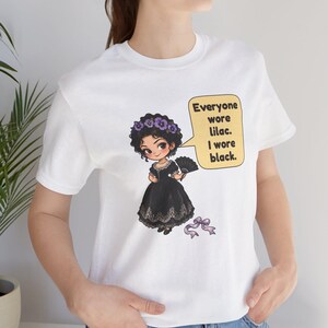 May include: A white t-shirt featuring a graphic of a cartoon character in a black dress with a purple floral crown and a hand fan. A speech bubble contains the text: Everyone wore lilac. I wore black. A small purple ribbon is positioned below the figure.