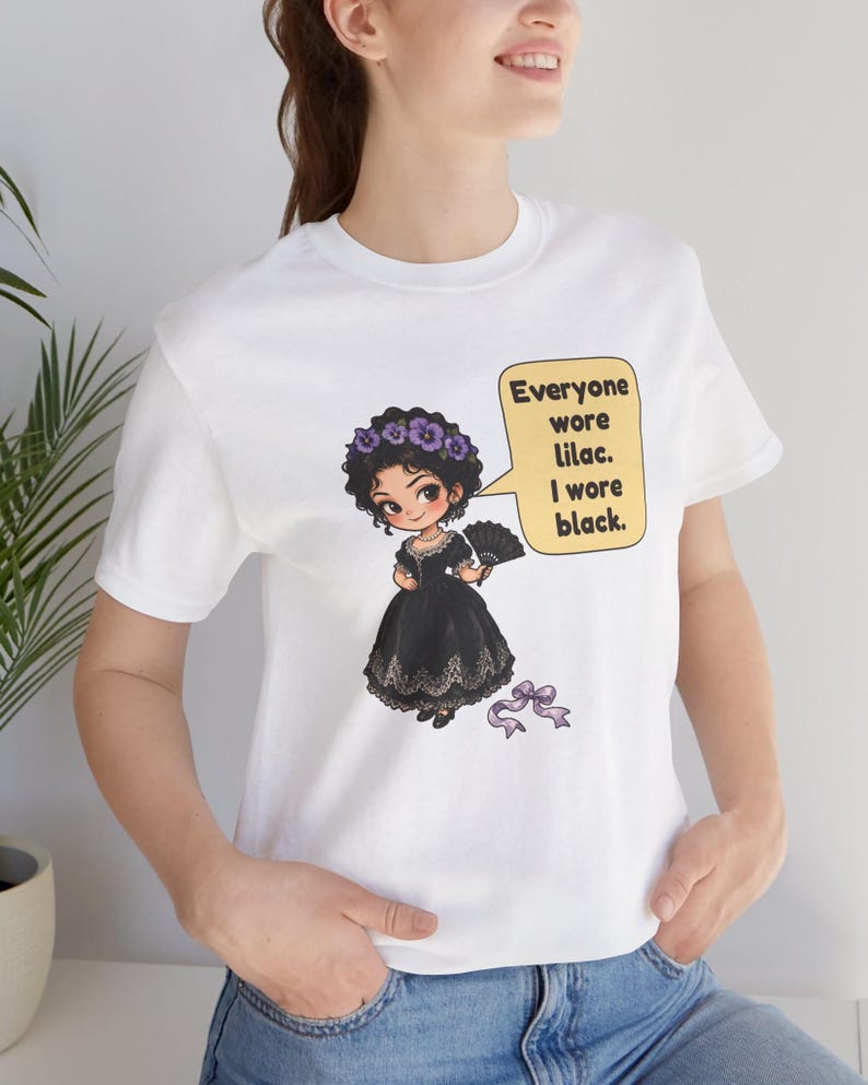 May include: A white t-shirt featuring a graphic of a cartoon character in a black dress with a purple floral crown and a hand fan. A speech bubble contains the text: Everyone wore lilac. I wore black. A small purple ribbon is positioned below the figure.