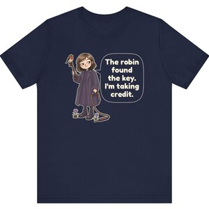 May include: A navy blue t-shirt featuring a graphic of a person in a dark coat holding a large key with a robin perched on it. A speech bubble contains the text: The robin found the key. I'm taking credit. The design includes small purple flowers.