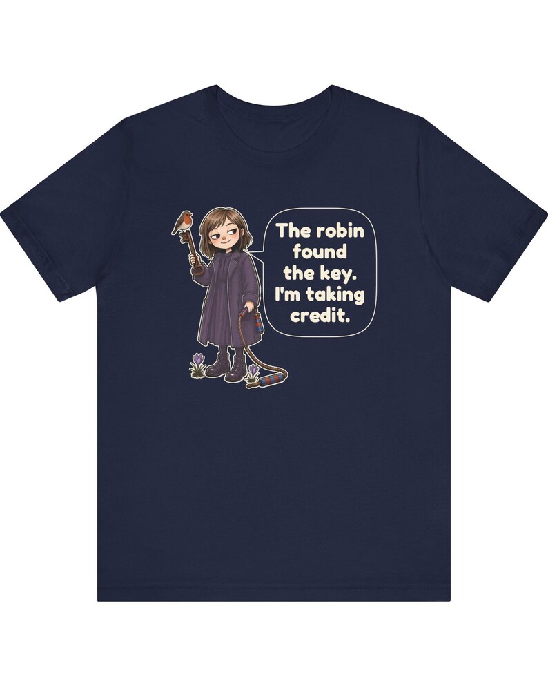 May include: A navy blue t-shirt featuring a graphic of a person in a dark coat holding a large key with a robin perched on it. A speech bubble contains the text: The robin found the key. I'm taking credit. The design includes small purple flowers.
