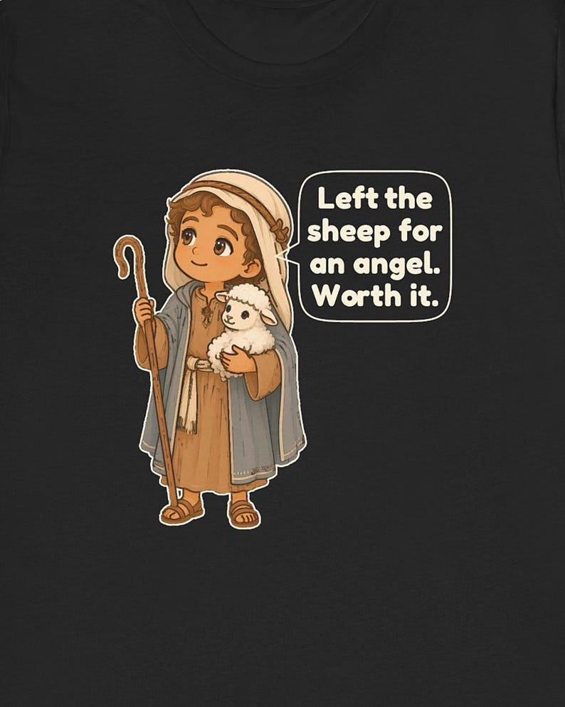 May include: A black t-shirt featuring a cartoon illustration of a shepherd wearing a tan tunic and blue cloak, holding a wooden staff and a small white sheep. A speech bubble contains the text: Left the sheep for an angel. Worth it.