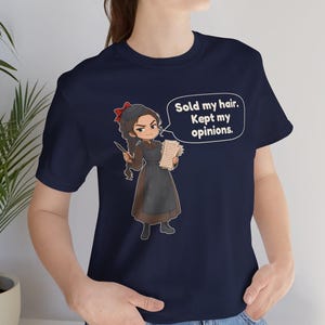 May include: A navy blue short-sleeved t-shirt featuring a cartoon illustration of a person with a braid and red hair bow, holding a quill and papers. A speech bubble contains the text: Sold my hair. Kept my opinions.