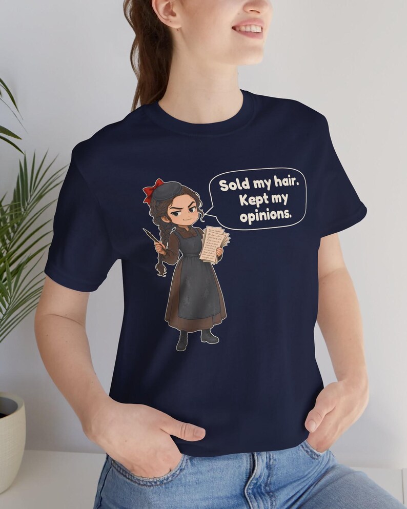May include: A navy blue short-sleeved t-shirt featuring a cartoon illustration of a person with a braid and red hair bow, holding a quill and papers. A speech bubble contains the text: Sold my hair. Kept my opinions.