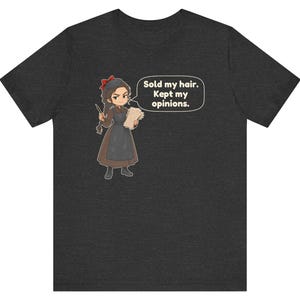 May include: A dark heather gray t-shirt featuring a cartoon illustration of a person with braided hair wearing a brown dress and black apron. The character holds a quill and paper, with a speech bubble containing the text: Sold my hair. Kept my opinions.