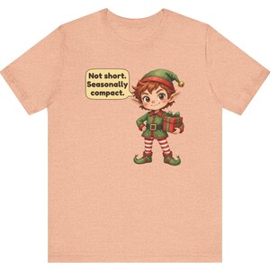 May include: A peach-colored short-sleeve t-shirt featuring a graphic of a cartoon elf wearing a green outfit and red and white striped leggings, holding a gift box. A speech bubble next to the elf contains the text: Not short. Seasonally compact.