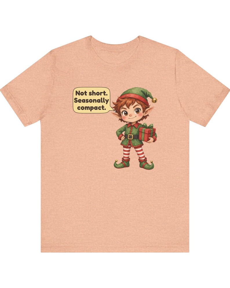 May include: A peach-colored short-sleeve t-shirt featuring a graphic of a cartoon elf wearing a green outfit and red and white striped leggings, holding a gift box. A speech bubble next to the elf contains the text: Not short. Seasonally compact.