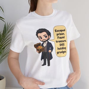May include: A white t-shirt featuring a cartoon illustration of a man in a dark blue coat holding a treasure chest. A yellow speech bubble contains the text: Escaped prison. Found treasure. Still holding grudges.