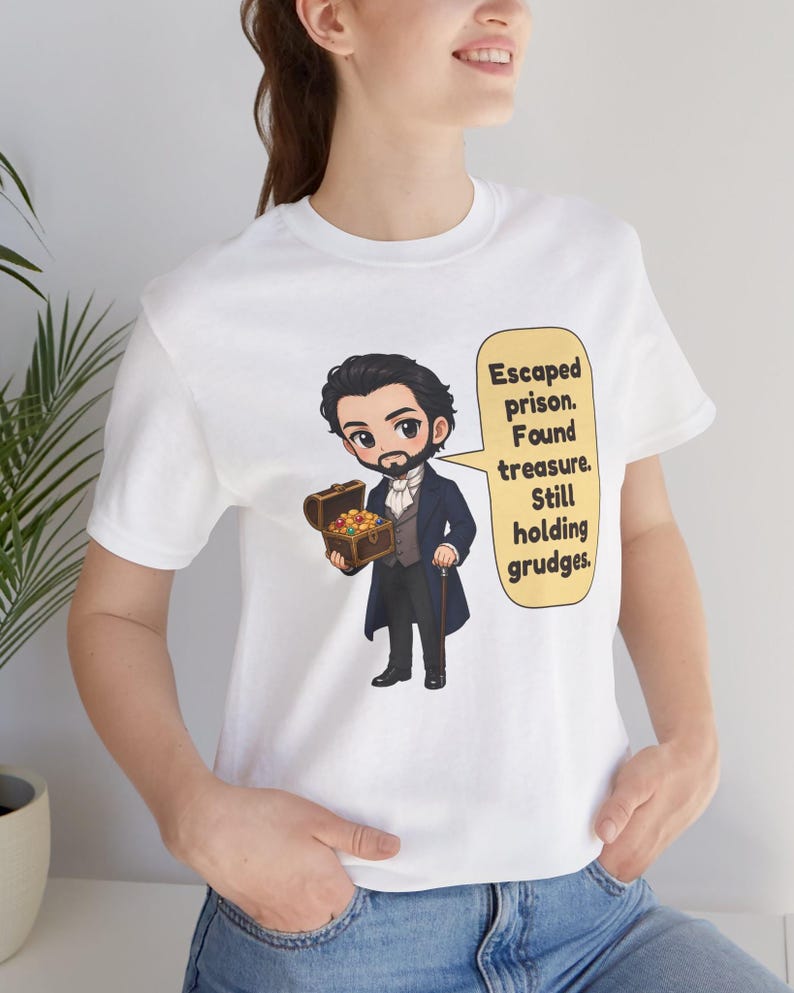 May include: A white t-shirt featuring a cartoon illustration of a man in a dark blue coat holding a treasure chest. A yellow speech bubble contains the text: Escaped prison. Found treasure. Still holding grudges.