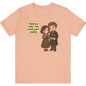 May include: A peach-colored t-shirt featuring a cartoon illustration of a couple holding a gift box. A speech bubble above the female character contains the text: Sold my hair. You sold your watch. The characters are dressed in brown and dark clothing.