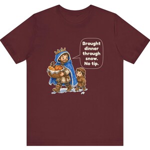 May include: A maroon t-shirt featuring a graphic of a king in a blue cape carrying a basket of bread with a child, accompanied by a speech bubble that reads: Brought dinner through snow. No tip.