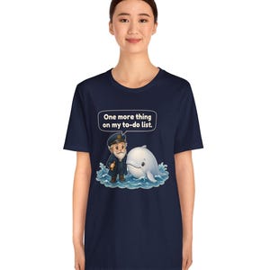 May include: A navy blue short-sleeve t-shirt featuring a graphic of a sea captain standing next to a white whale in blue water. Above the characters, a speech bubble contains the text: One more thing on my to-do list.