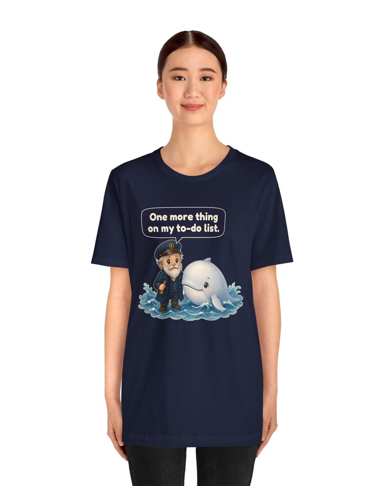 May include: A navy blue short-sleeve t-shirt featuring a graphic of a sea captain standing next to a white whale in blue water. Above the characters, a speech bubble contains the text: One more thing on my to-do list.