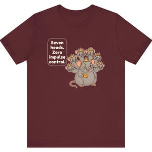 May include: A maroon short-sleeve t-shirt featuring a graphic of a seven-headed mouse wearing small gold crowns and holding a piece of yellow cheese. The text "Seven heads. Zero impulse control." is printed in a white speech bubble.