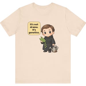 May include: A light beige t-shirt featuring a cartoon illustration of a monk holding a pea pod and a watering can. A speech bubble next to the monk contains the text: It's not drama. It's genetics. The design is centered on the front of the shirt.