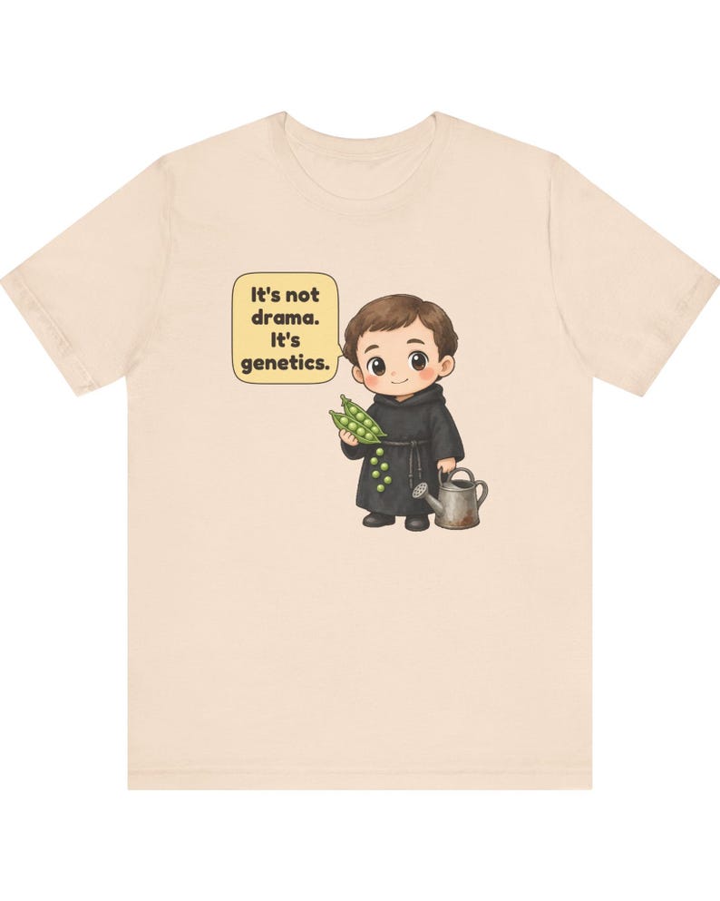 May include: A light beige t-shirt featuring a cartoon illustration of a monk holding a pea pod and a watering can. A speech bubble next to the monk contains the text: It's not drama. It's genetics. The design is centered on the front of the shirt.