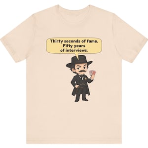 Pu&ograve; includere: Una t-shirt beige chiaro con un'illustrazione a cartoni animati di una figura con i baffi, cappello e abito nero che tiene in mano delle carte da gioco. Un fumetto sopra la figura contiene il testo: Thirty seconds of fame. Fifty years of interviews.