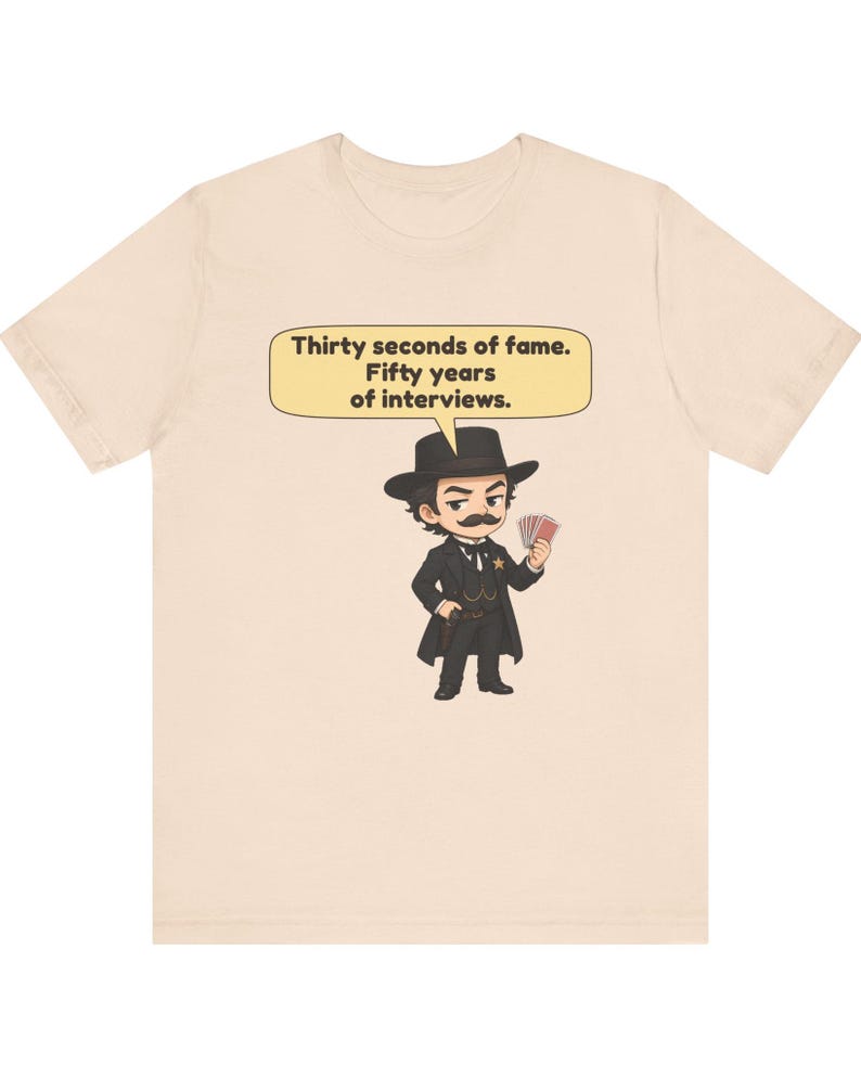 Pu&ograve; includere: Una t-shirt beige chiaro con un'illustrazione a cartoni animati di una figura con i baffi, cappello e abito nero che tiene in mano delle carte da gioco. Un fumetto sopra la figura contiene il testo: Thirty seconds of fame. Fifty years of interviews.