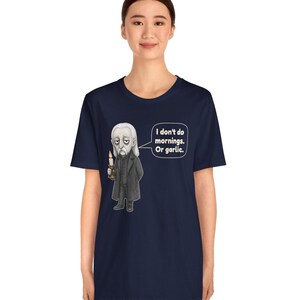 May include: A navy blue short-sleeve t-shirt featuring a graphic of a pale, long-haired character in a black coat holding a lit candle. A speech bubble next to the character contains the text: I don't do mornings. Or garlic.