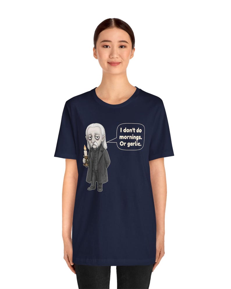 May include: A navy blue short-sleeve t-shirt featuring a graphic of a pale, long-haired character in a black coat holding a lit candle. A speech bubble next to the character contains the text: I don't do mornings. Or garlic.