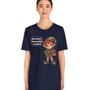May include: A navy blue t-shirt featuring a cartoon elf wearing a green and red outfit with a matching hat, holding a red gift box. A speech bubble next to the elf contains the text: Not short. Seasonally compact.