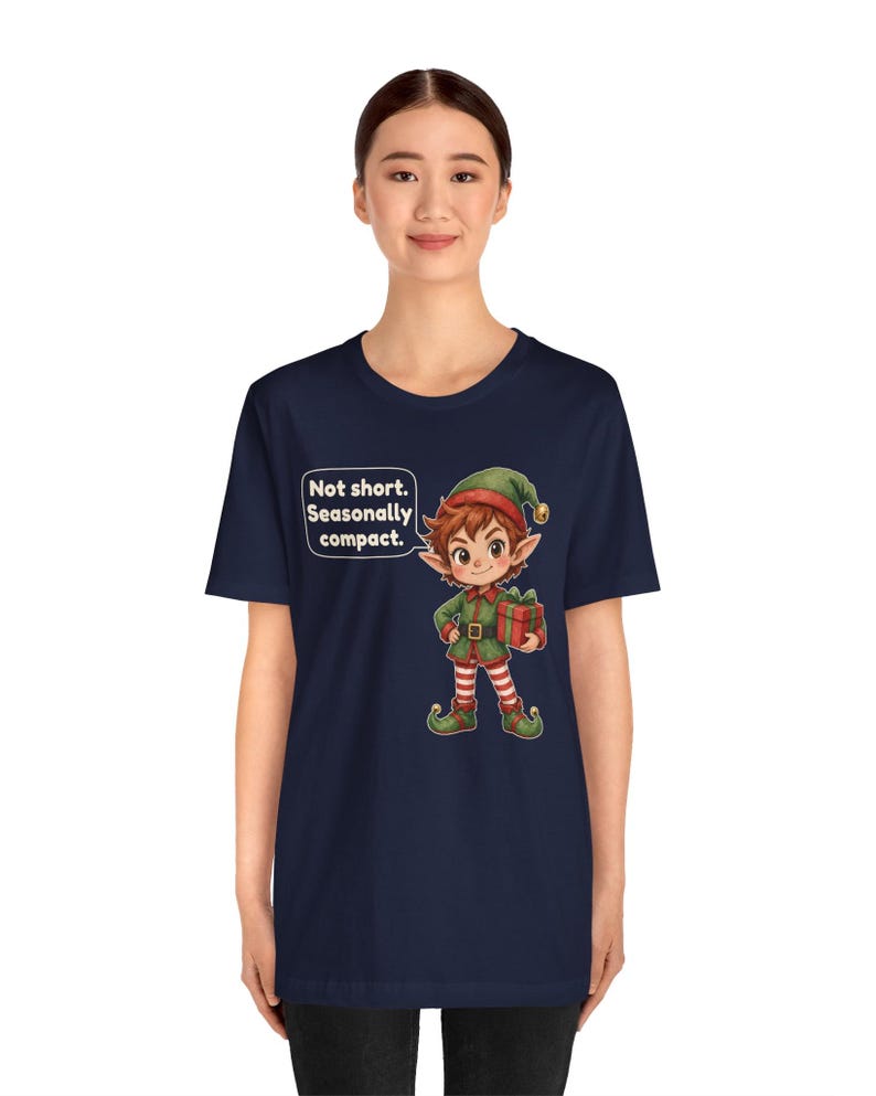 May include: A navy blue t-shirt featuring a cartoon elf wearing a green and red outfit with a matching hat, holding a red gift box. A speech bubble next to the elf contains the text: Not short. Seasonally compact.