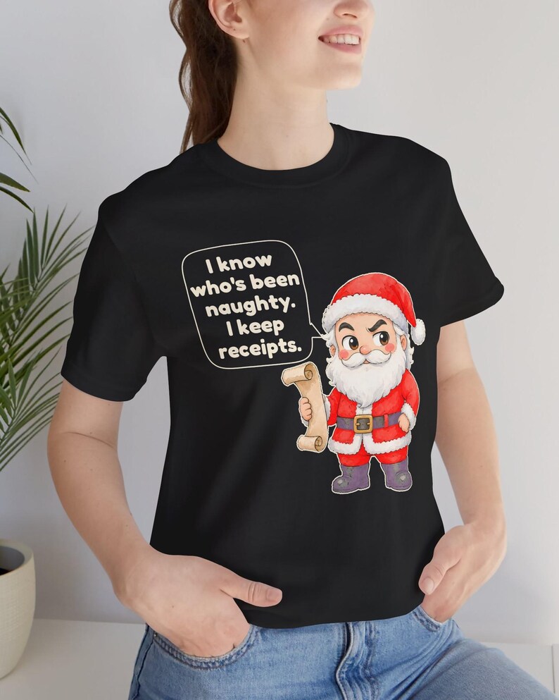 May include: A black t-shirt featuring a cartoon illustration of Santa Claus holding a scroll. The text in a white speech bubble reads: I know who's been naughty. I keep receipts. Santa wears a red suit with white trim and dark boots.
