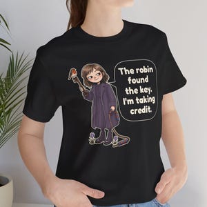 May include: A black short-sleeve t-shirt featuring a cartoon illustration of a person in a dark coat holding a large key with a robin perched on it. The text reads, The robin found the key. I'm taking credit. The design includes small purple flowers.