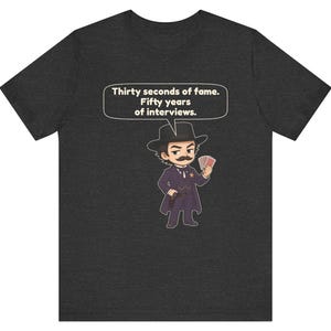 Pu&ograve; includere: Una t-shirt grigio scuro m&eacute;lange con un'illustrazione in stile cartoon di un personaggio con baffi, cappello da cowboy, cappotto e distintivo a stella, che tiene in mano delle carte. Un fumetto sopra riporta il testo: Thirty seconds of fame. Fifty years of interviews.