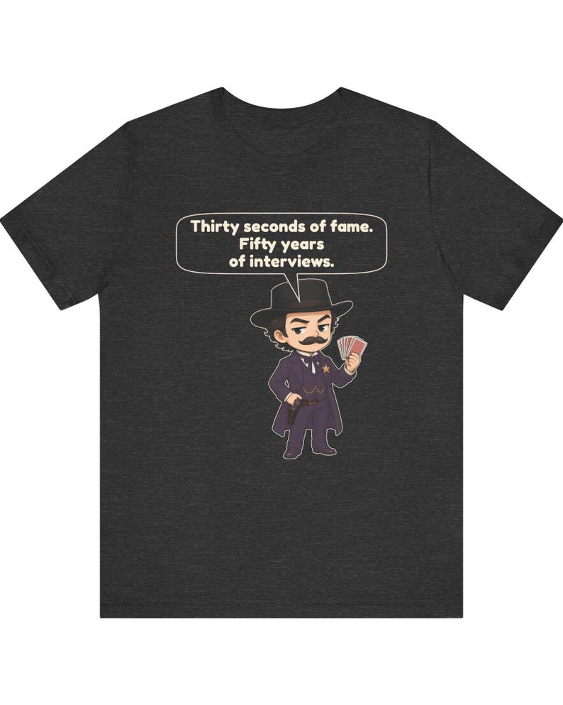 Pu&ograve; includere: Una t-shirt grigio scuro m&eacute;lange con un'illustrazione in stile cartoon di un personaggio con baffi, cappello da cowboy, cappotto e distintivo a stella, che tiene in mano delle carte. Un fumetto sopra riporta il testo: Thirty seconds of fame. Fifty years of interviews.