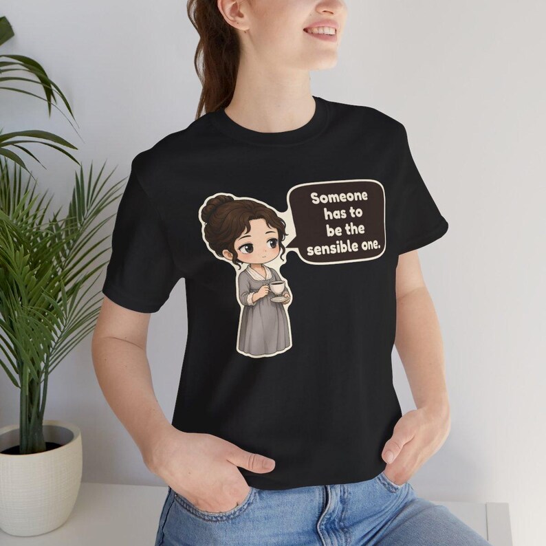 May include: A black short-sleeve t-shirt featuring a cartoon illustration of a person in a gray dress holding a teacup. A speech bubble next to the character contains the text, Someone has to be the sensible one. The design is centered on the chest.